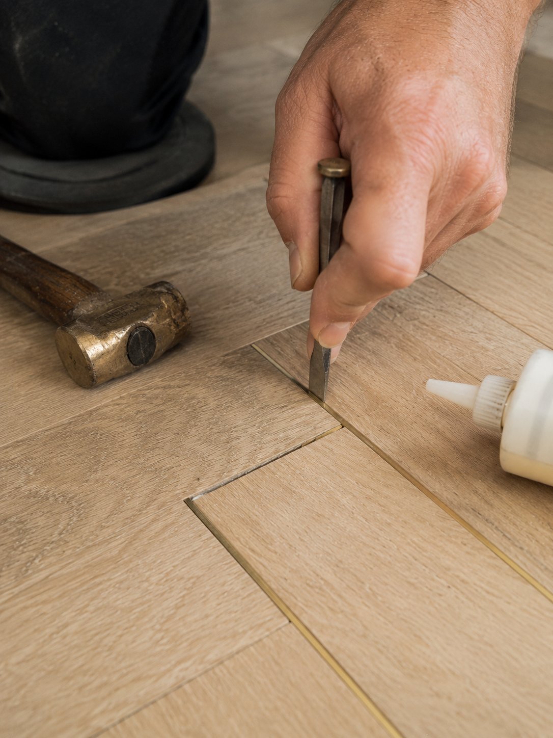 A fitter setting a brass inlay strip into the channel between two oak panels with a small chisel, brass mallet and glue applicator alongside
