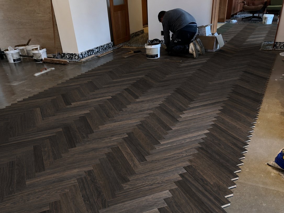 A Timberzone fitter kneeling at the far end of a room mid-installation — dark smoked oak herringbone advancing diagonally across the floor, paint buckets and tools alongside