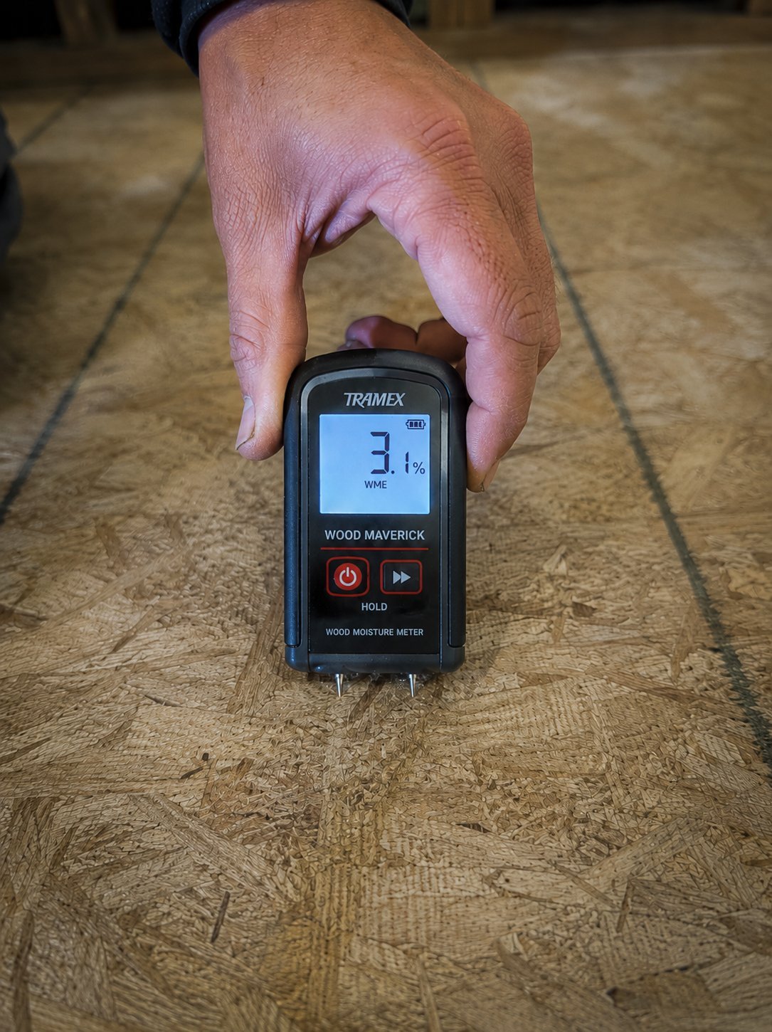 A Tramex Wood Maverick moisture meter showing 3.1% WME pressed to an OSB subfloor — moisture check before installation
